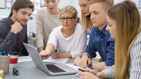 Ältere Schülerinnen und Schüler bekommen von einer Lehrerin etwas auf einem Laptop gezeigt.
